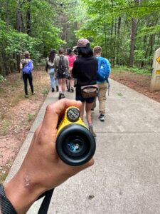 A hand holds a monocular in front of a group of people on a forested trail.