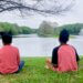 Two children sit on grass and look at a lake with a fountain.