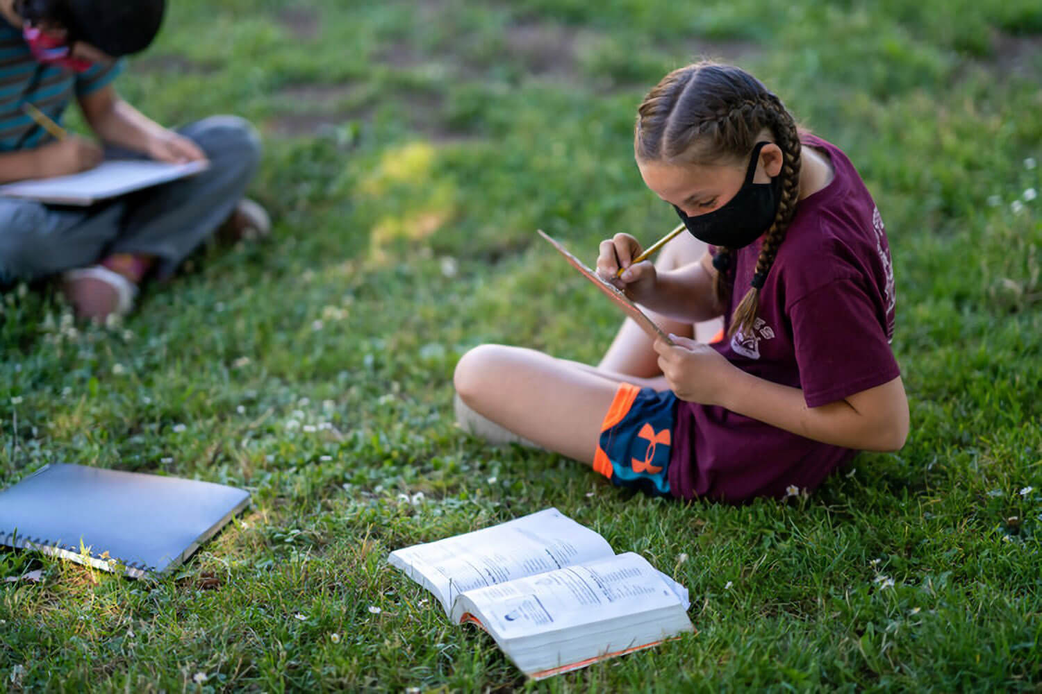 Young girl sitting cross-legged in grass working with a book and notepad.